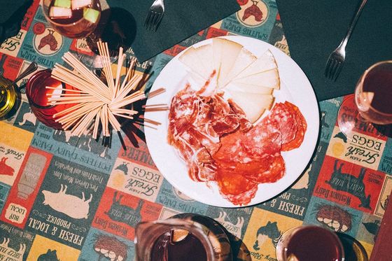 A table with a plate of cured meats and cheese, surrounded by drinks, napkins, and forks on a colorful, patterned tablecloth.