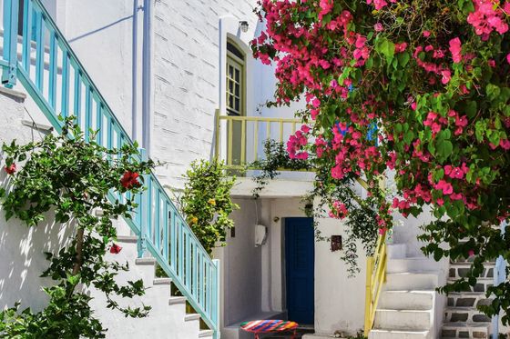 A charming narrow alley with white buildings, colorful railings, vibrant pink flowers, and a small table in a Mediterranean village.