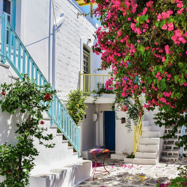 A charming narrow alley with white buildings, colorful railings, vibrant pink flowers, and a small table in a Mediterranean village.