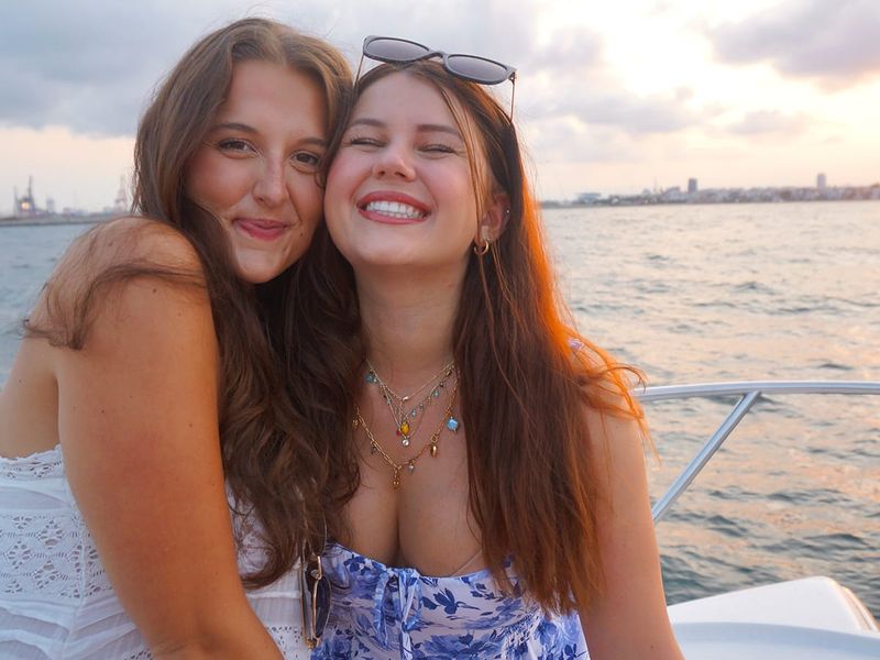 Two smiling women on a boat at sunset, with one wearing sunglasses and floral dress, the other in white lace, against a sea and cloudy sky backdrop.
