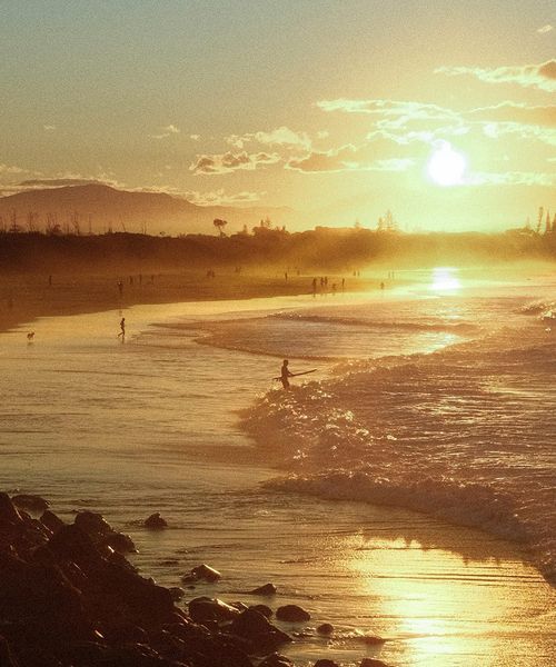 Sunset over a sandy beach with gentle waves, silhouetted people walking, and distant hills under a golden sky with scattered clouds.