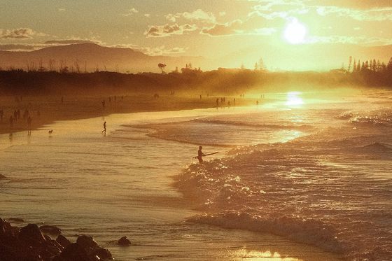 Sunset over a sandy beach with gentle waves, silhouetted people walking, and distant hills under a golden sky with scattered clouds.