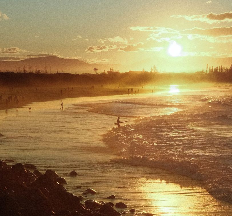Sunset over a sandy beach with gentle waves, silhouetted people walking, and distant hills under a golden sky with scattered clouds.