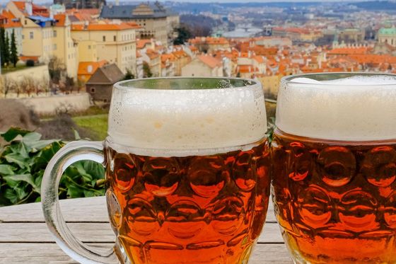 Two mugs of amber beer with foam sit on a wooden table, overlooking a European cityscape with historic buildings and a cathedral in the background.