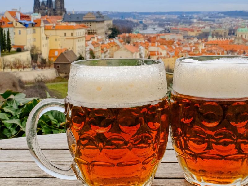 Two mugs of amber beer with foam sit on a wooden table, overlooking a European cityscape with historic buildings and a cathedral in the background.
