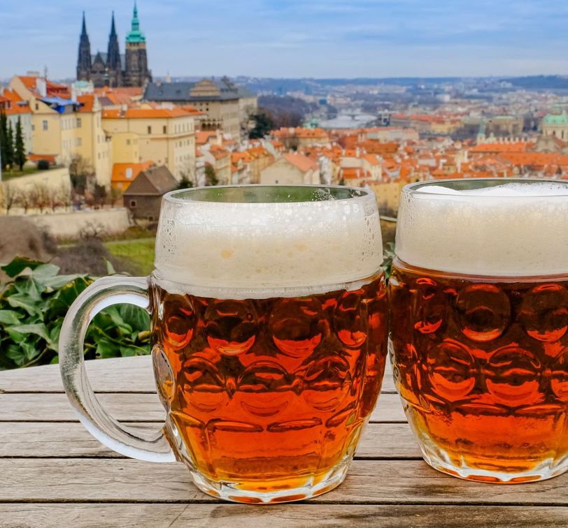 Two mugs of amber beer with foam sit on a wooden table, overlooking a European cityscape with historic buildings and a cathedral in the background.