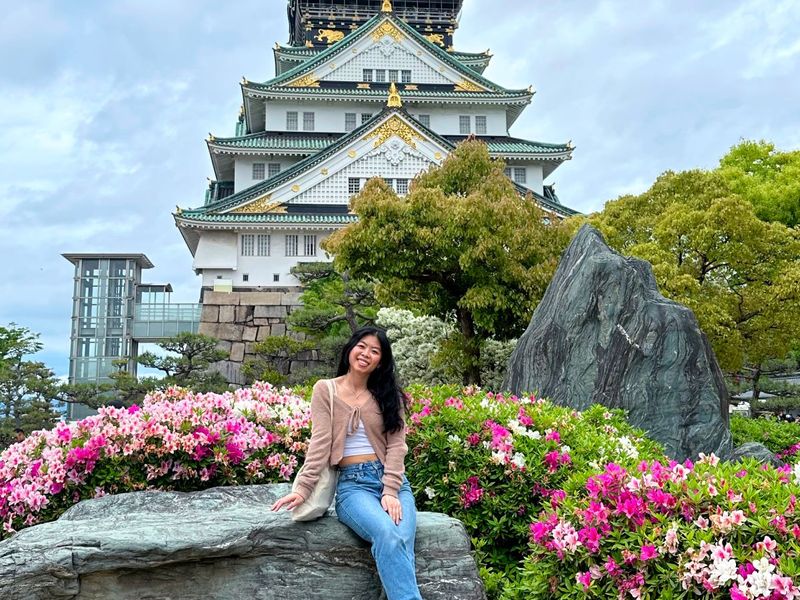 Person sitting on a rock, smiling, surrounded by pink and white flowers, with a large traditional Japanese building in the background.