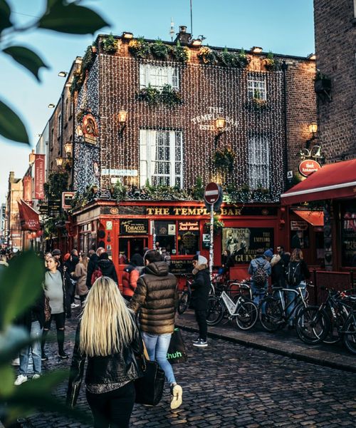 People walking outside The Temple Bar in Dublin, Ireland, with festive lights and bicycles nearby on a cobblestone street.