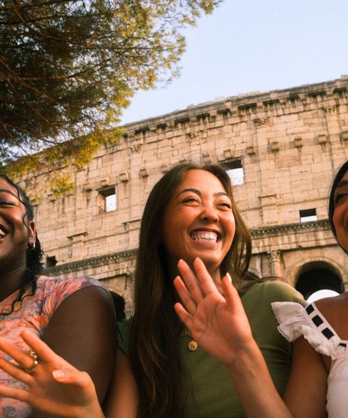 Three friends laughing and smiling in front of the Colosseum, with trees overhead and a clear blue sky.