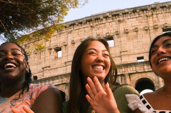 Three friends laughing and smiling in front of the Colosseum, with trees overhead and a clear blue sky.