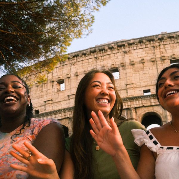 Three friends laughing and smiling in front of the Colosseum, with trees overhead and a clear blue sky.