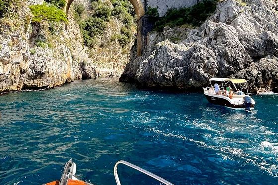 A boat approaches a rocky coastline with an arched stone bridge. Another boat is nearby on the vibrant blue water beneath lush cliffs.