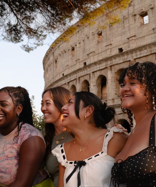 Four women smiling and laughing in front of the Colosseum, with sunlight casting a warm glow on them and the ancient structure.