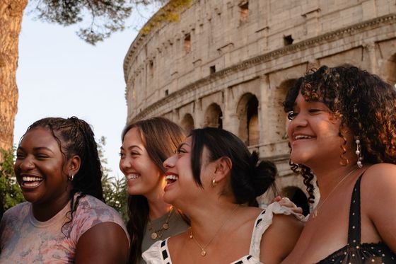 Four women smiling and laughing in front of the Colosseum, with sunlight casting a warm glow on them and the ancient structure.