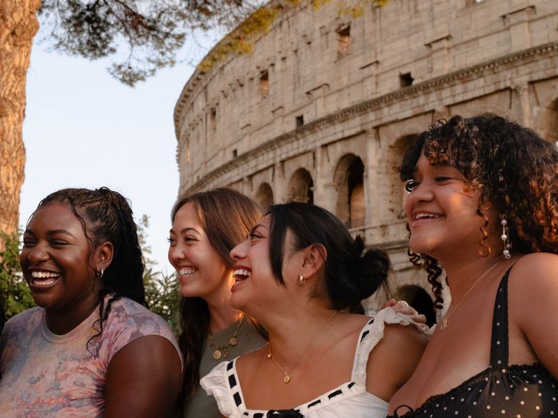 Four women smiling and laughing in front of the Colosseum, with sunlight casting a warm glow on them and the ancient structure.