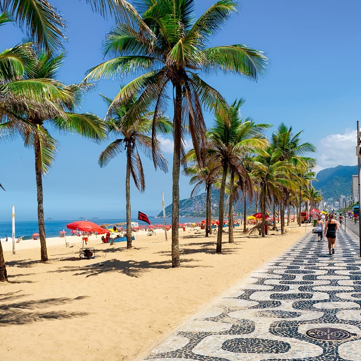 Palm-lined beach with red umbrellas and people relaxing. Patterned stone walkway on the right under a clear blue sky.