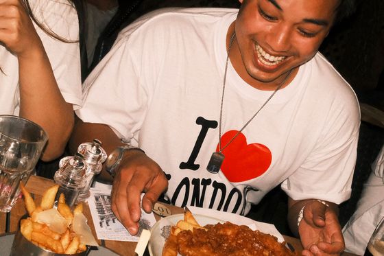 A person smiling at a plate of fish and chips, wearing a white "I ♥️ London" shirt, seated at a table with fries and a burger.