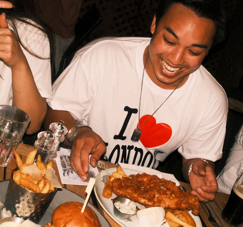 Smiling person wearing an "I ♥ London" shirt, seated at a table with fish and chips, a burger, and drinks.