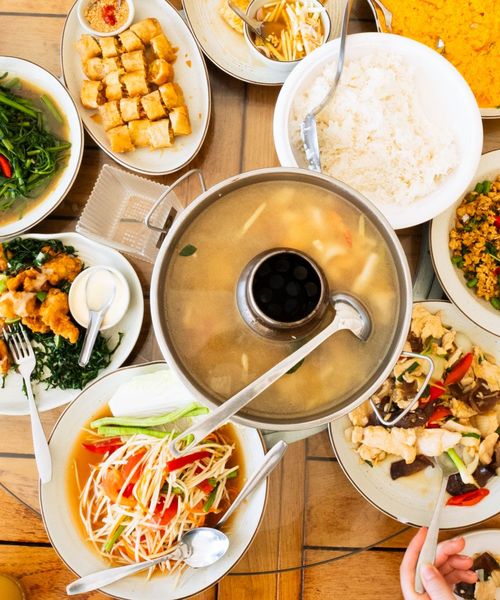 Assorted Asian dishes including soup, rice, stir-fried vegetables, papaya salad, and crispy rolls arranged on a wooden table.