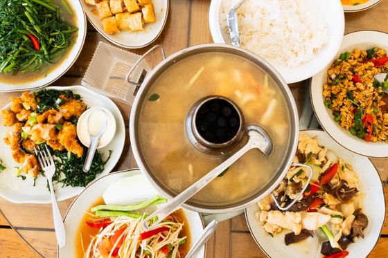 Assorted Asian dishes including soup, rice, stir-fried vegetables, papaya salad, and crispy rolls arranged on a wooden table.