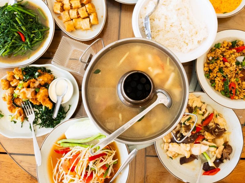 Assorted Asian dishes including soup, rice, stir-fried vegetables, papaya salad, and crispy rolls arranged on a wooden table.