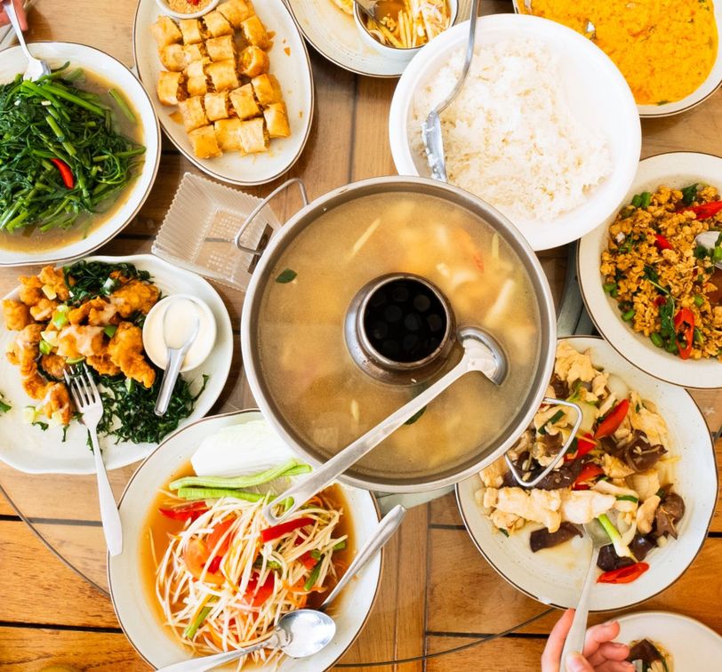 Assorted Asian dishes including soup, rice, stir-fried vegetables, papaya salad, and crispy rolls arranged on a wooden table.