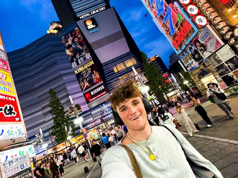 A person wearing headphones takes a selfie in a bustling city street at night, surrounded by colorful signs and a large hotel building.