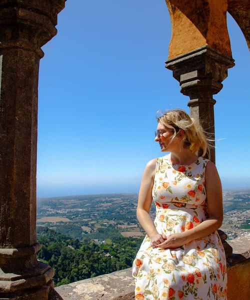 Woman in floral dress sits on a stone ledge, surrounded by arches, overlooking a scenic landscape under a clear blue sky.