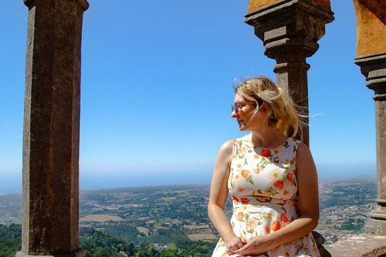 Woman in floral dress sits on a stone ledge, surrounded by arches, overlooking a scenic landscape under a clear blue sky.
