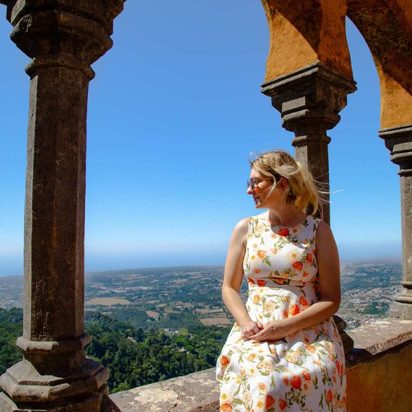 Woman in floral dress sits on a stone ledge, surrounded by arches, overlooking a scenic landscape under a clear blue sky.