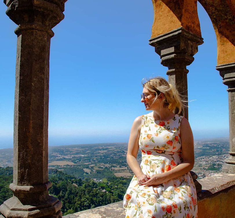 Woman in floral dress sits on a stone ledge, surrounded by arches, overlooking a scenic landscape under a clear blue sky.