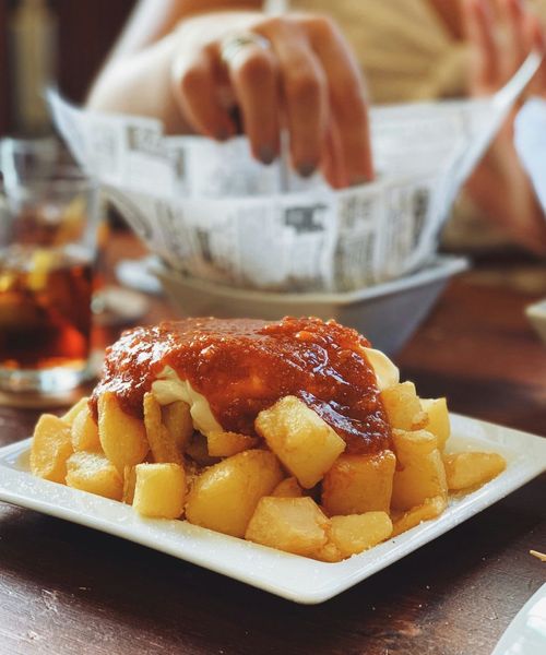 Crispy potatoes topped with a red sauce on a plate, with a hand reaching for food in the background. A drink is visible on the table.