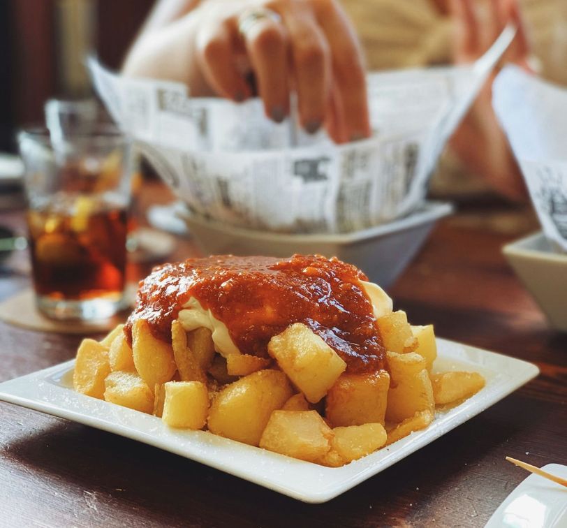 Crispy potatoes topped with a red sauce on a plate, with a hand reaching for food in the background. A drink is visible on the table.