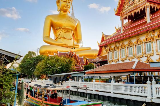 A large golden Buddha statue beside ornate temple buildings with a colorful boat on a canal in the foreground.
