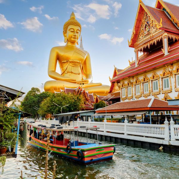 A large golden Buddha statue beside ornate temple buildings with a colorful boat on a canal in the foreground.