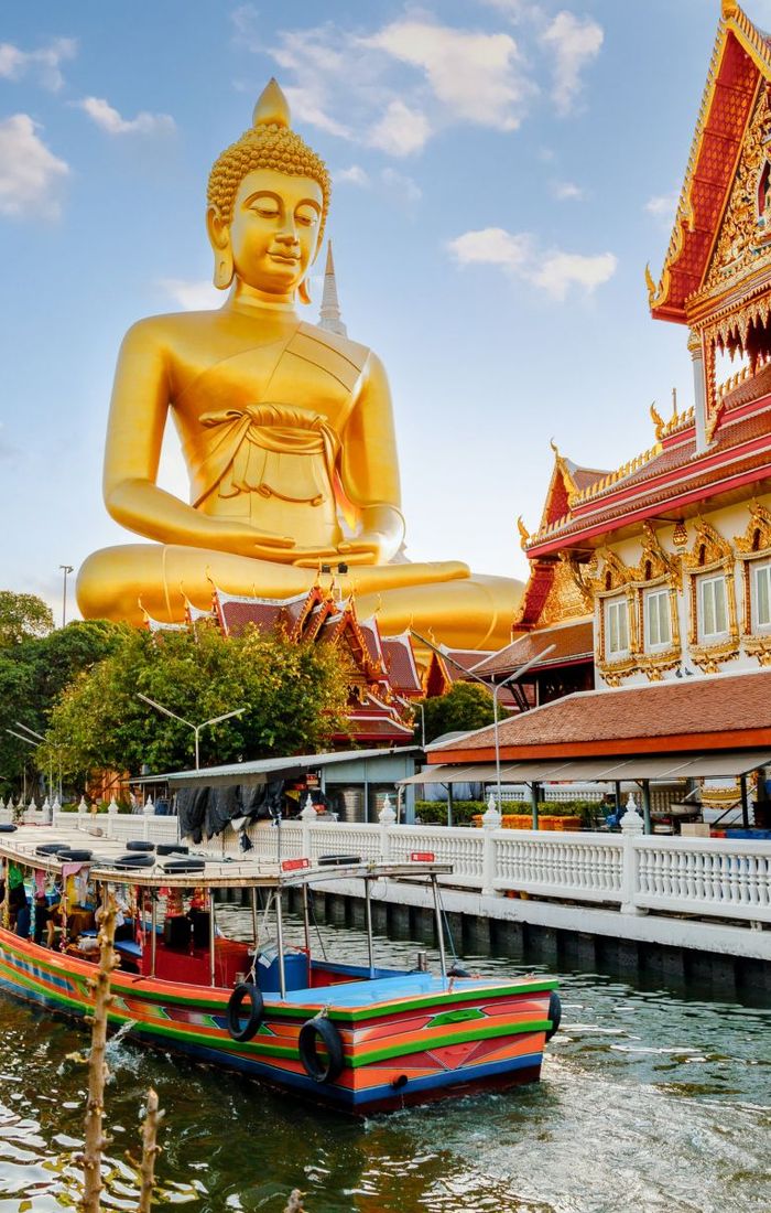 A large golden Buddha statue beside ornate temple buildings with a colorful boat on a canal in the foreground.
