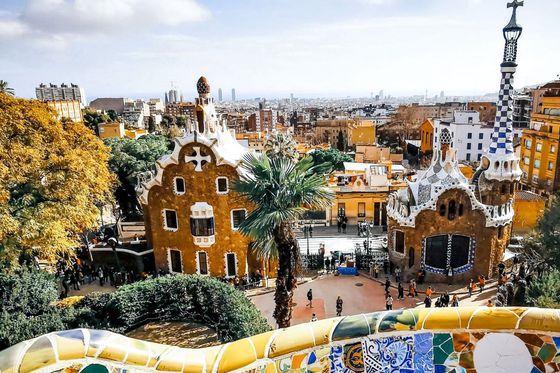 Park Güell in Barcelona, with colorful mosaic tiles and whimsical architecture, set against a cityscape under a partly cloudy blue sky.