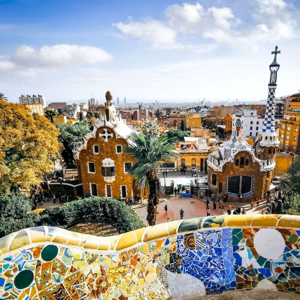 Park Güell in Barcelona, with colorful mosaic tiles and whimsical architecture, set against a cityscape under a partly cloudy blue sky.