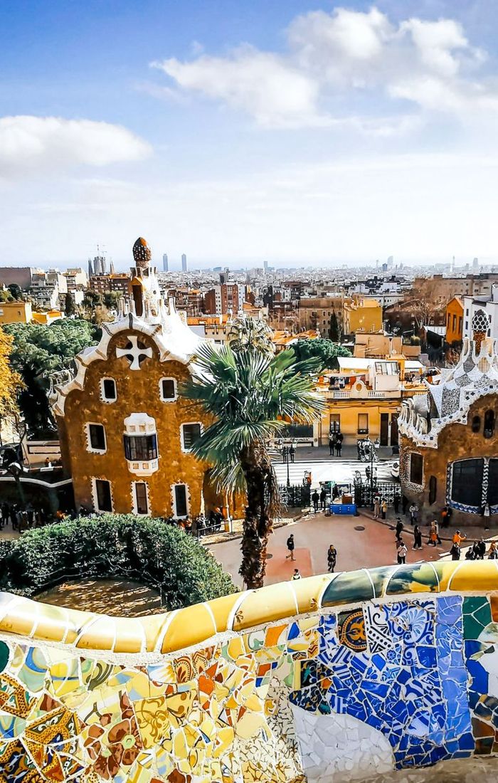 Park Güell in Barcelona, with colorful mosaic tiles and whimsical architecture, set against a cityscape under a partly cloudy blue sky.