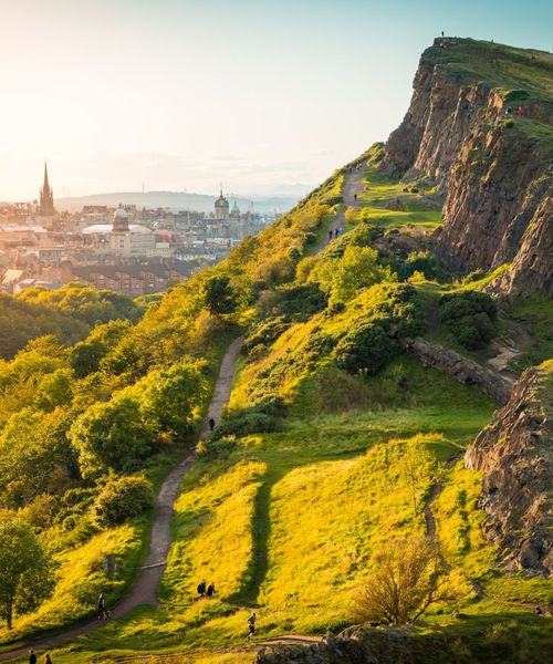A scenic view of a sunlit green hillside with a winding path, overlooking a cityscape with spires and church towers in the background.
