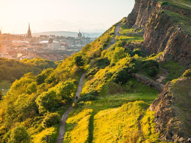 A scenic view of a sunlit green hillside with a winding path, overlooking a cityscape with spires and church towers in the background.
