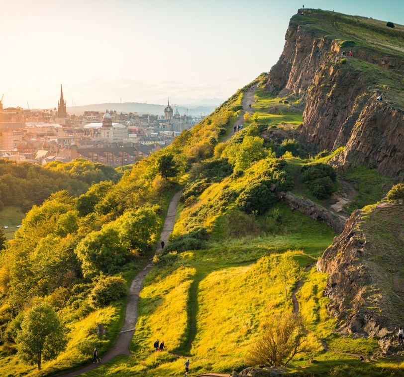 A scenic view of a sunlit green hillside with a winding path, overlooking a cityscape with spires and church towers in the background.