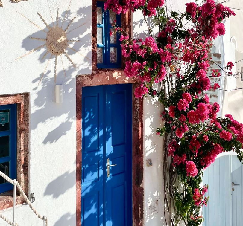 White wall with a vibrant blue door, adorned by climbing pink bougainvillea. A decorative sun hangs nearby, casting shadows.
