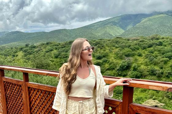 Woman in light outfit stands on a wooden deck, enjoying a lush green mountain view under a partly cloudy sky.