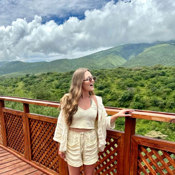Woman in light outfit stands on a wooden deck, enjoying a lush green mountain view under a partly cloudy sky.