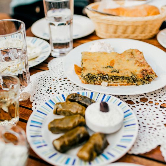 A table with spinach pie, grape leaf rolls with a dollop of cream, glasses of water, and bread in a basket on a crochet tablecloth.