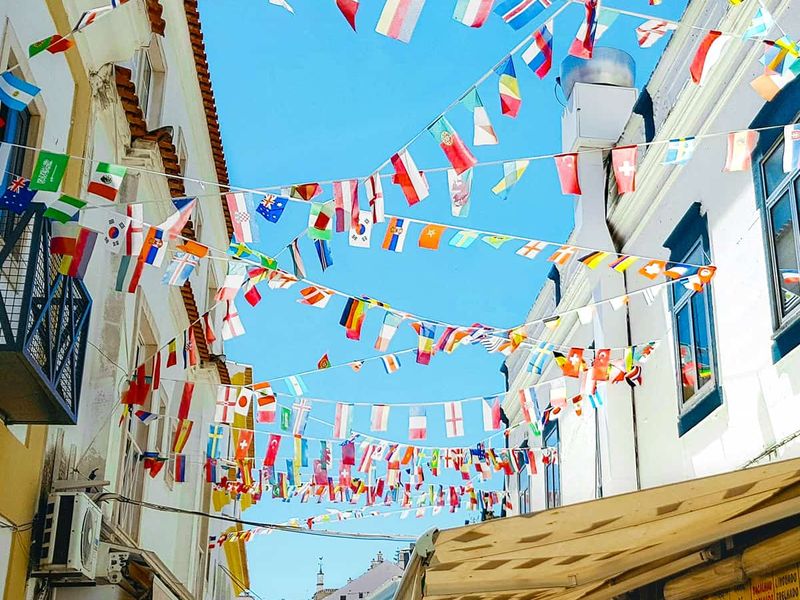Narrow street with colorful international flags strung between buildings under a clear blue sky.