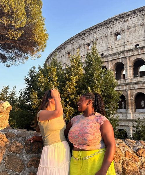 Two women smiling at each other beside a stone wall with the Colosseum in the background, under a clear blue sky.