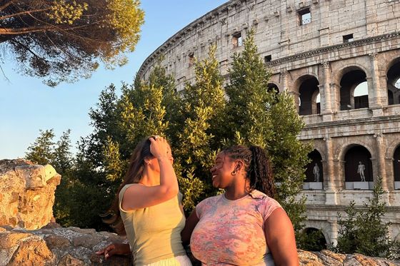 Two women smiling at each other beside a stone wall with the Colosseum in the background, under a clear blue sky.