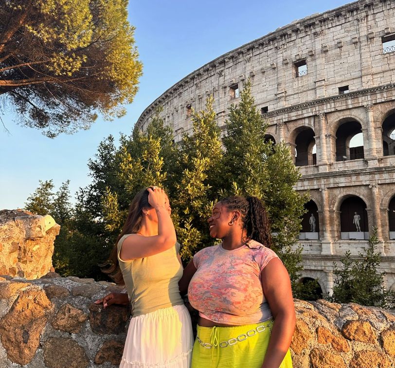 Two women smiling at each other beside a stone wall with the Colosseum in the background, under a clear blue sky.